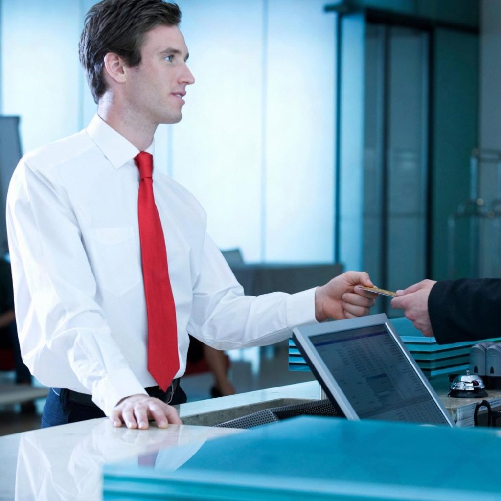 Front desk agent in a white shirt and red tie handing a credit card back to a guest at a hotel reception counter